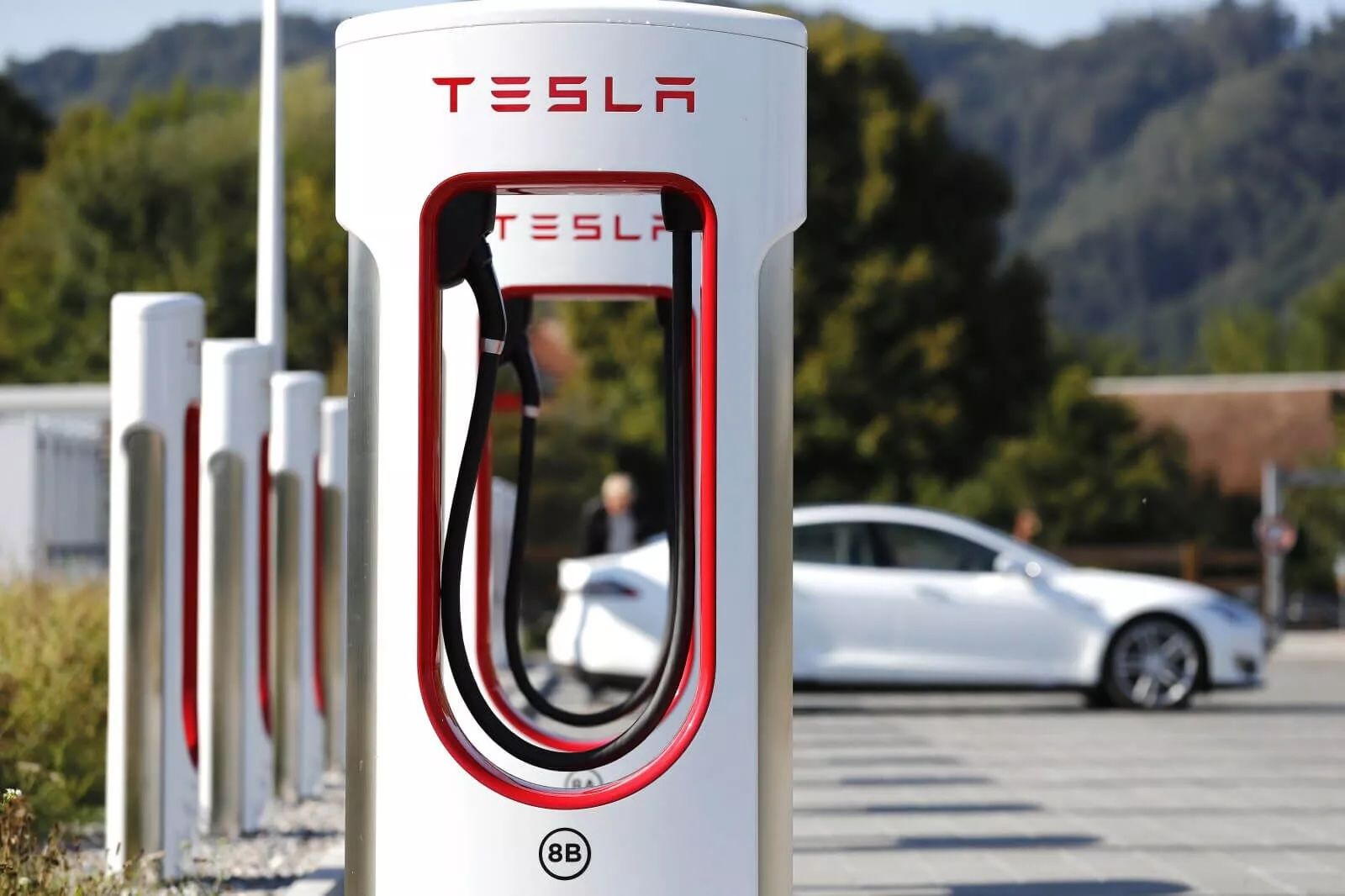 Station de recharge Tesla avec plusieurs chargeurs alignés, une voiture Tesla blanche garée à proximité et des arbres en arrière-plan.