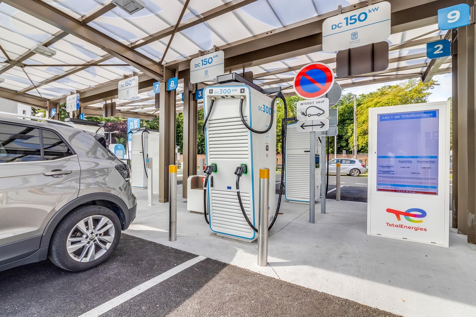 Borne de recharge pour véhicules électriques avec plusieurs unités de recharge, une voiture garée et la marque TotalEnergies sous une zone couverte.