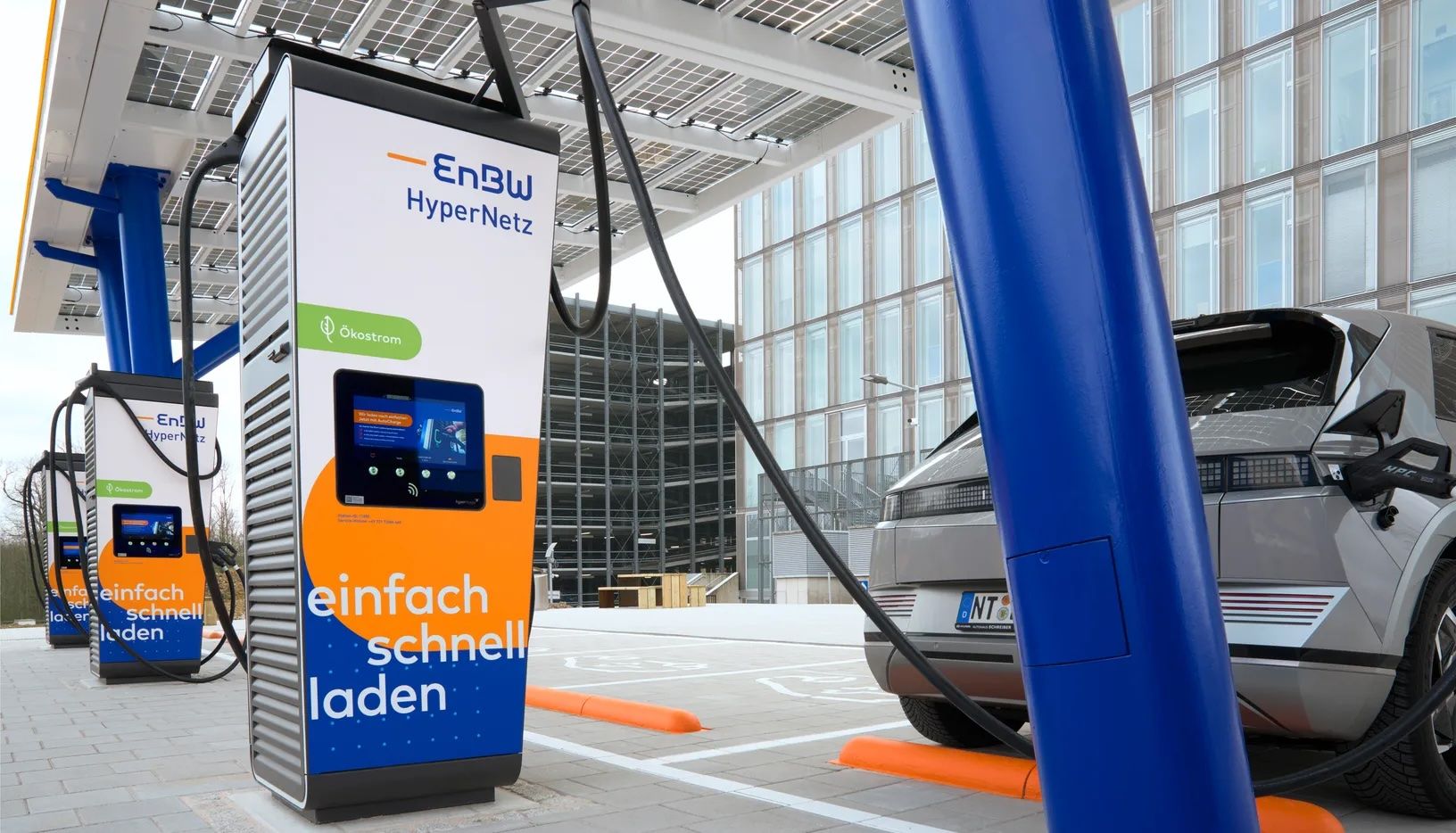 Ein Schnellladegerät von EnBW mit modernen orangefarbenen und blauen Mustern, angeschlossen an ein graues Elektrofahrzeug, das hinter einer blauen Säule vor einem großen, vollständig mit Glasfenstern bedeckten Gebäude geparkt ist.