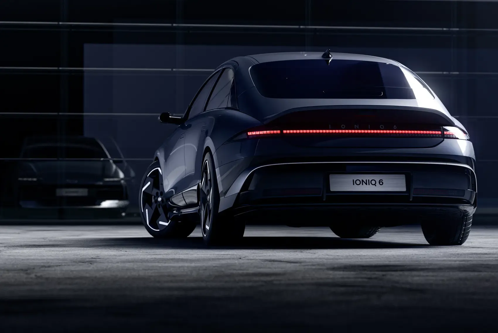 Elegante automóvil eléctrico Hyundai Ioniq 6 de color oscuro mostrado desde atrás, estacionado en un garaje poco iluminado con paredes de vidrio reflectante.