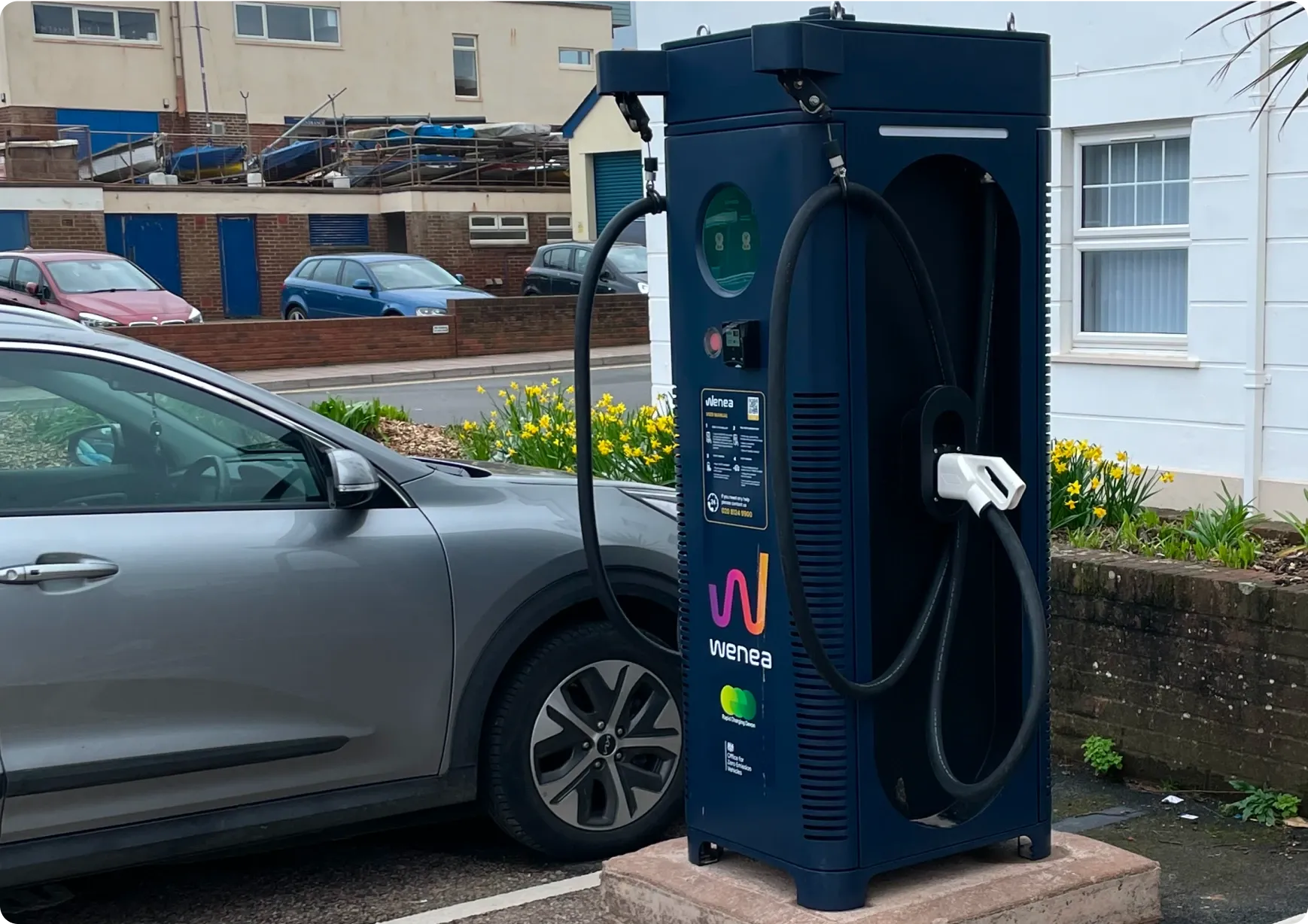 Estación de carga para vehículos eléctricos Wenea con cable integrado, junto a un coche gris aparcado. Cerca, flores amarillas y edificios residenciales.