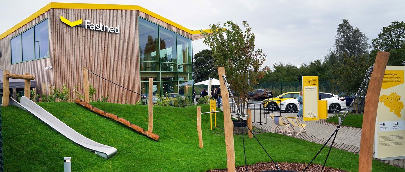 Una moderna stazione di ricarica Fastned con esterni in legno, circondata dal verde, uno scivolo e auto elettriche parcheggiate all'esterno.