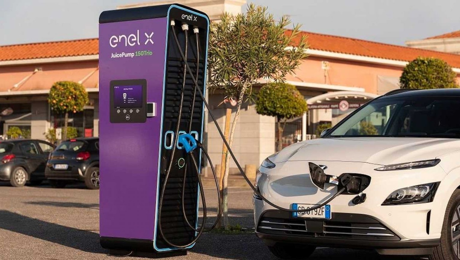 Ricarica di un veicolo elettrico presso una colonnina di ricarica viola Enel X in un parcheggio all'aperto soleggiato.