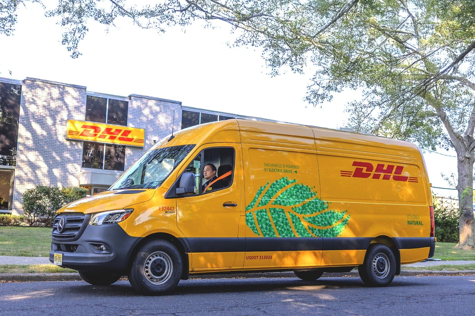 Furgone giallo della DHL con un disegno di foglie verdi, parcheggiato su una strada vicino a un edificio della DHL, con alberi sullo sfondo.