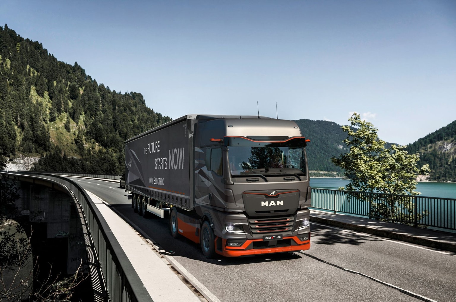 An electric MAN truck driving over a bridge with a lake and mountains in the background. Ein elektrischer MAN-LKW fährt über eine Brücke mit einem See und Bergen im Hintergrund