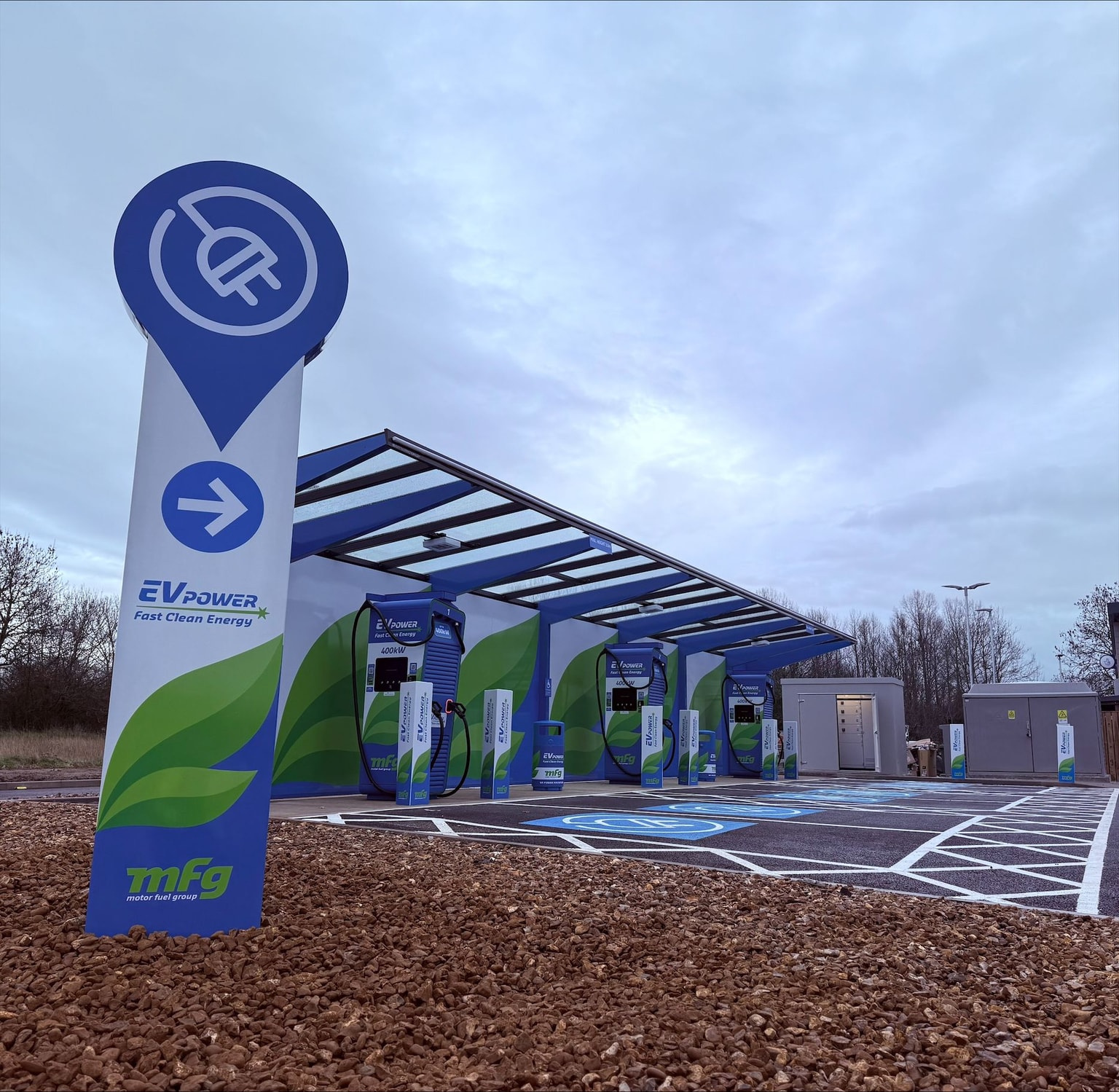 Electric vehicle charging station with multiple charging points under a canopy. Signage and branding visible. Overcast sky background.