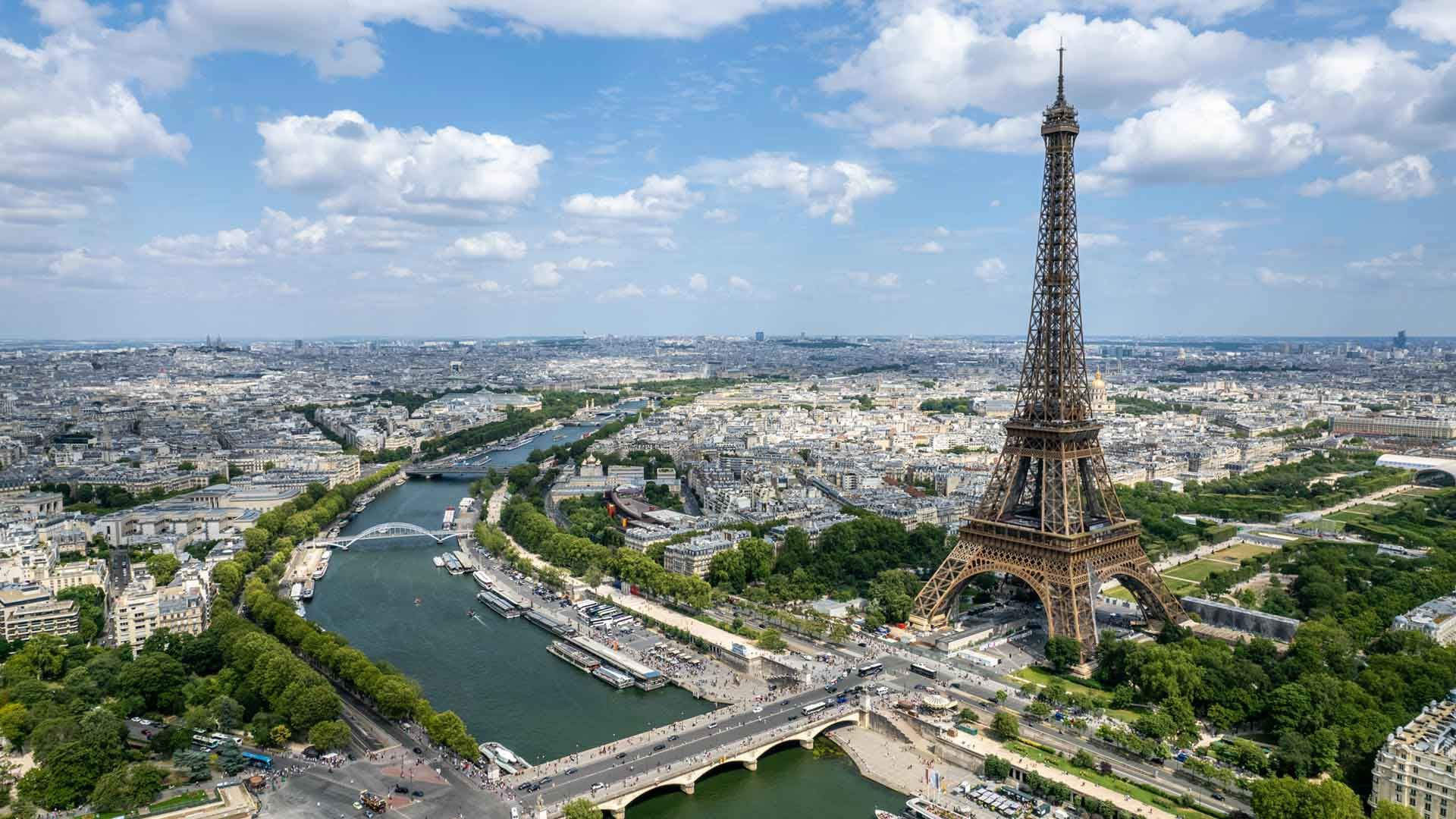 Vue aérienne de la Tour Eiffel à Paris, entourée par la Seine, les ponts et le paysage urbain sous un ciel partiellement nuageux.