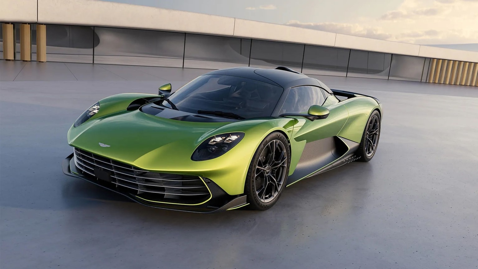 Ein Bild des Konzepts, ein grüner Aston Martin Valhalla, geparkt auf grauem Beton mit einem modernen Gebäude im Hintergrund und einem bewölkten Sonnenuntergangshimmel.