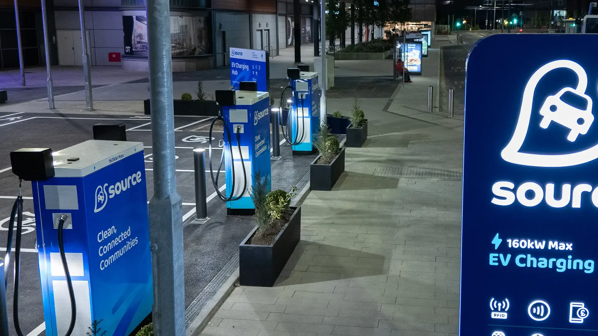 A row of electric vehicle charging stations illuminated at night, featuring blue signage and surrounded by small planters along a paved path.