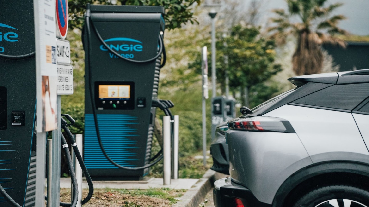 Voiture électrique garée à une borne de recharge extérieure, avec de la verdure et des arbres en arrière-plan.