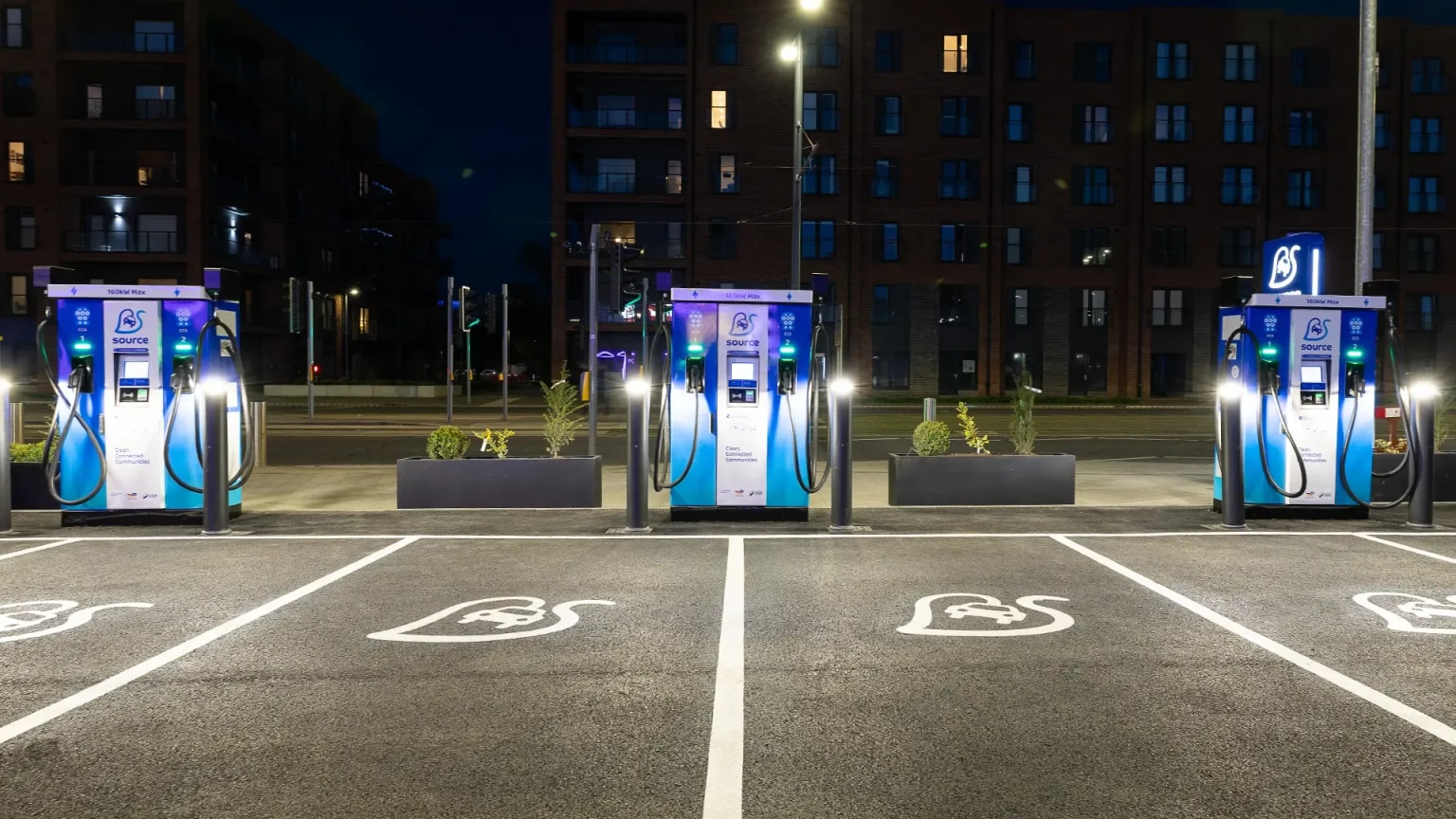 Three electric vehicle charging stations in a well-lit parking lot at night, with buildings in the background.