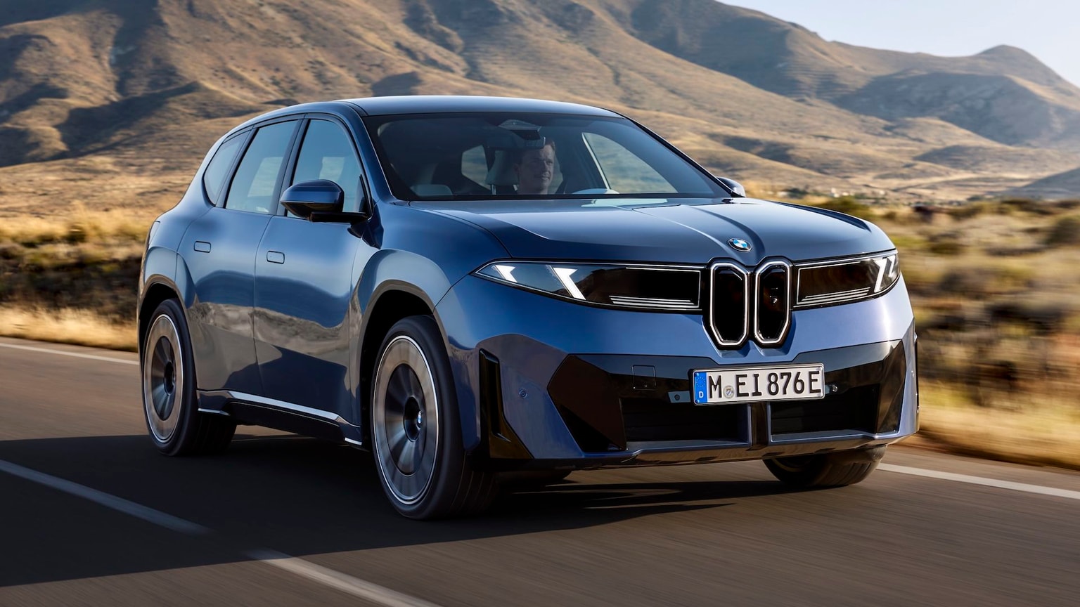 Ein Konzeptbild des BMW ix3, das Auto fährt auf einer Straße mit Gras und Bergen im Hintergrund.