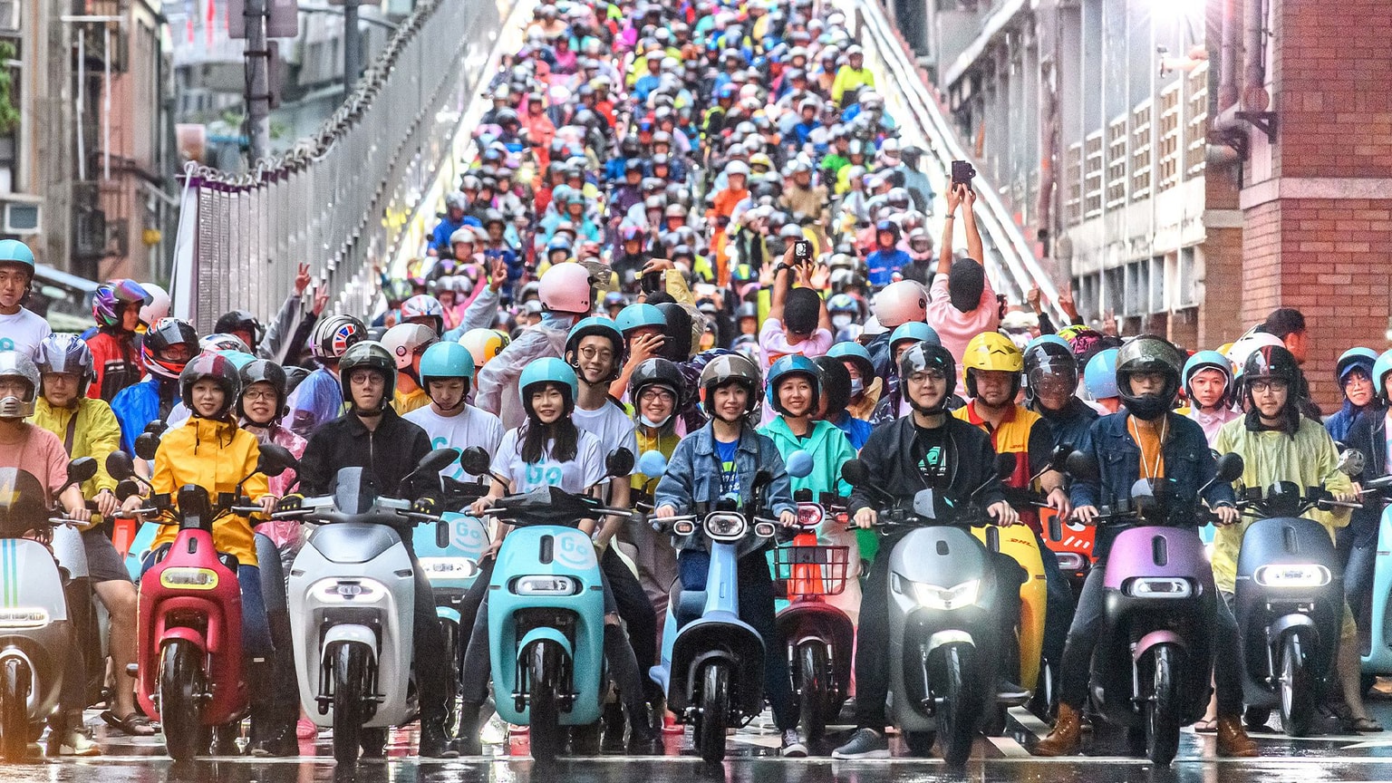 A large crowd of people on electric scooters, wearing helmets, gather on a rainy street at the base of a sloping bridge.