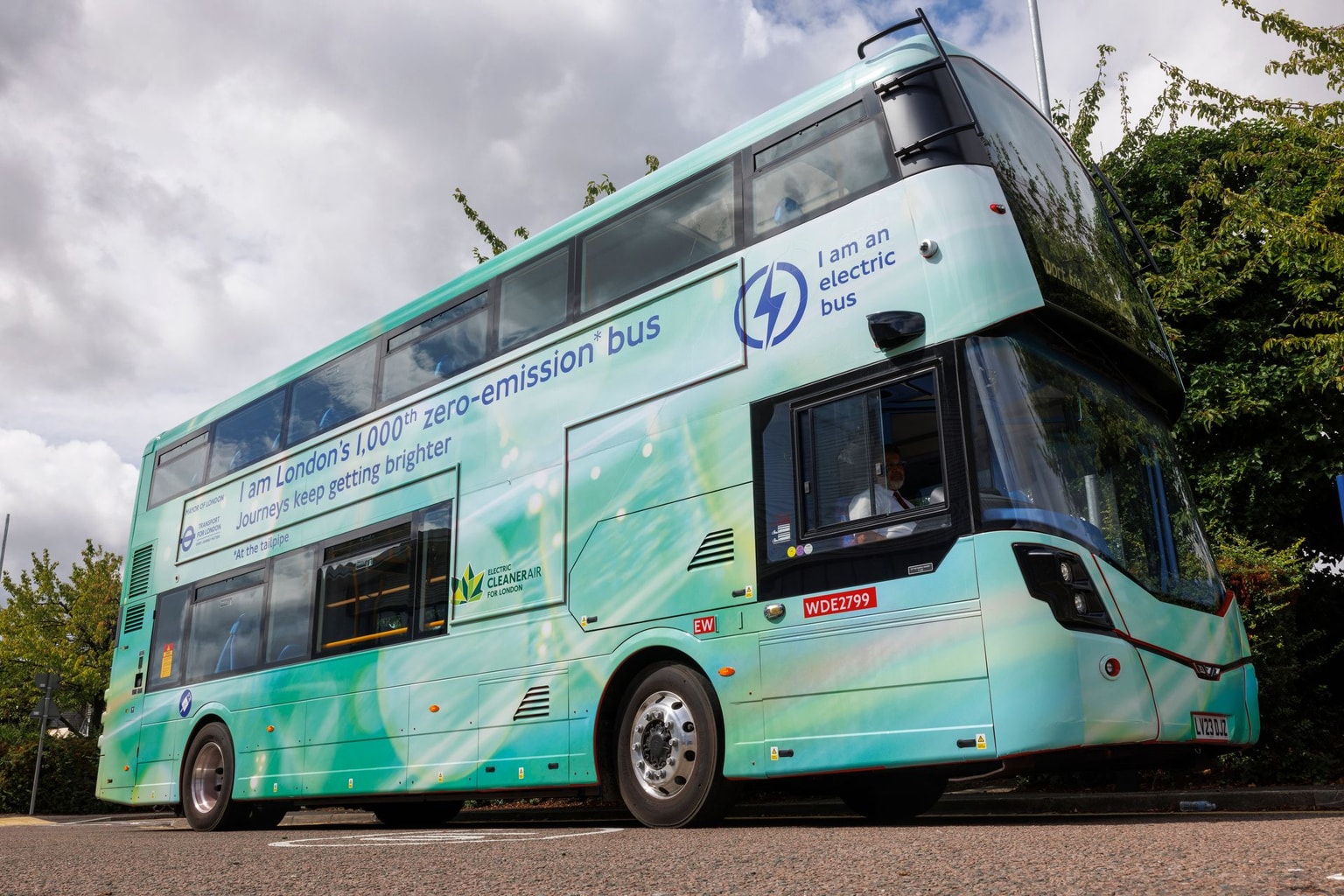 A light green double-decker electric bus on a road, promoting zero emissions, with cloudy sky and trees in the background.