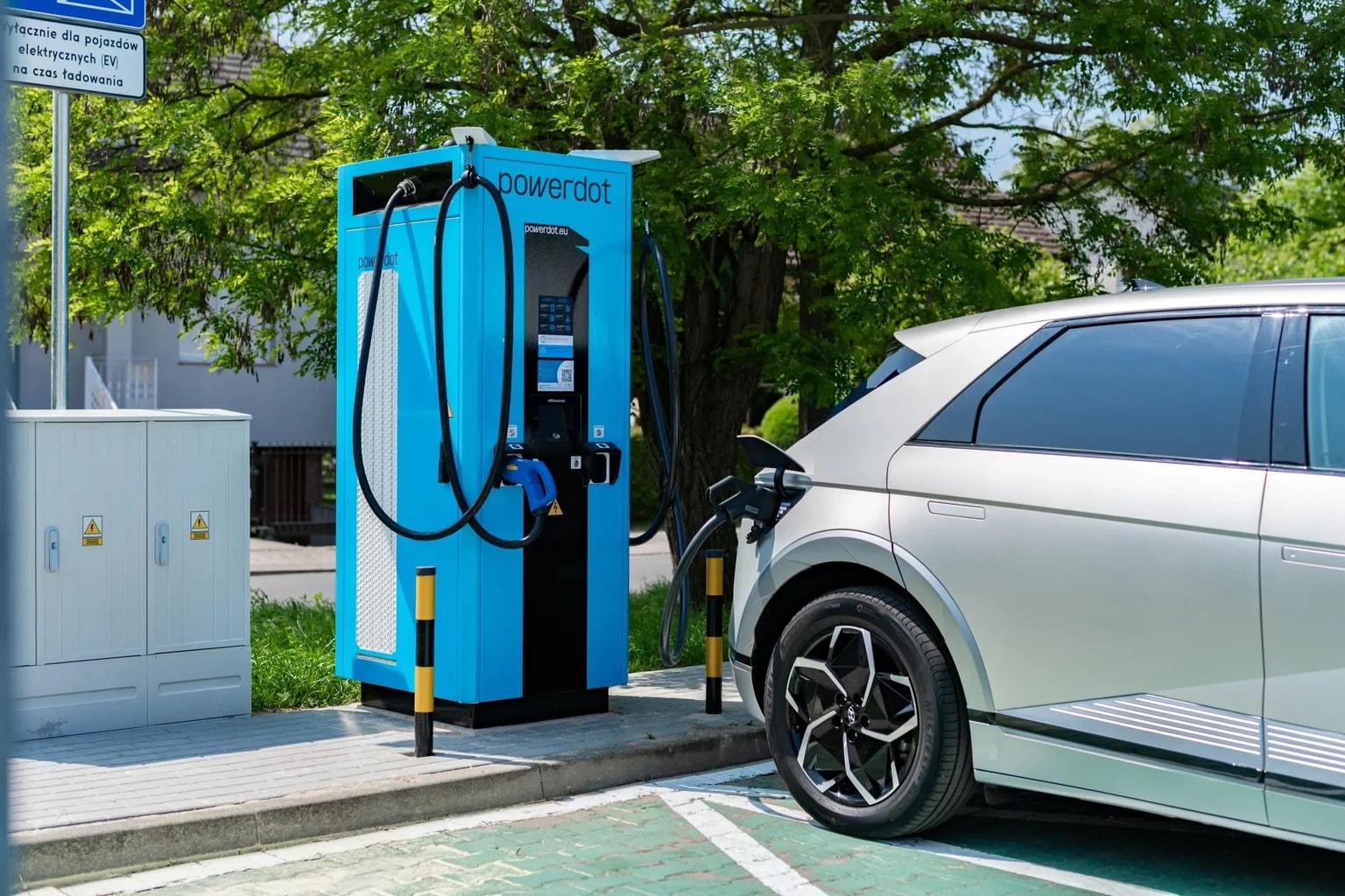 Recharge de voiture électrique sur une borne de recharge Powerdot bleue dans un cadre extérieur ensoleillé avec des arbres en arrière-plan.