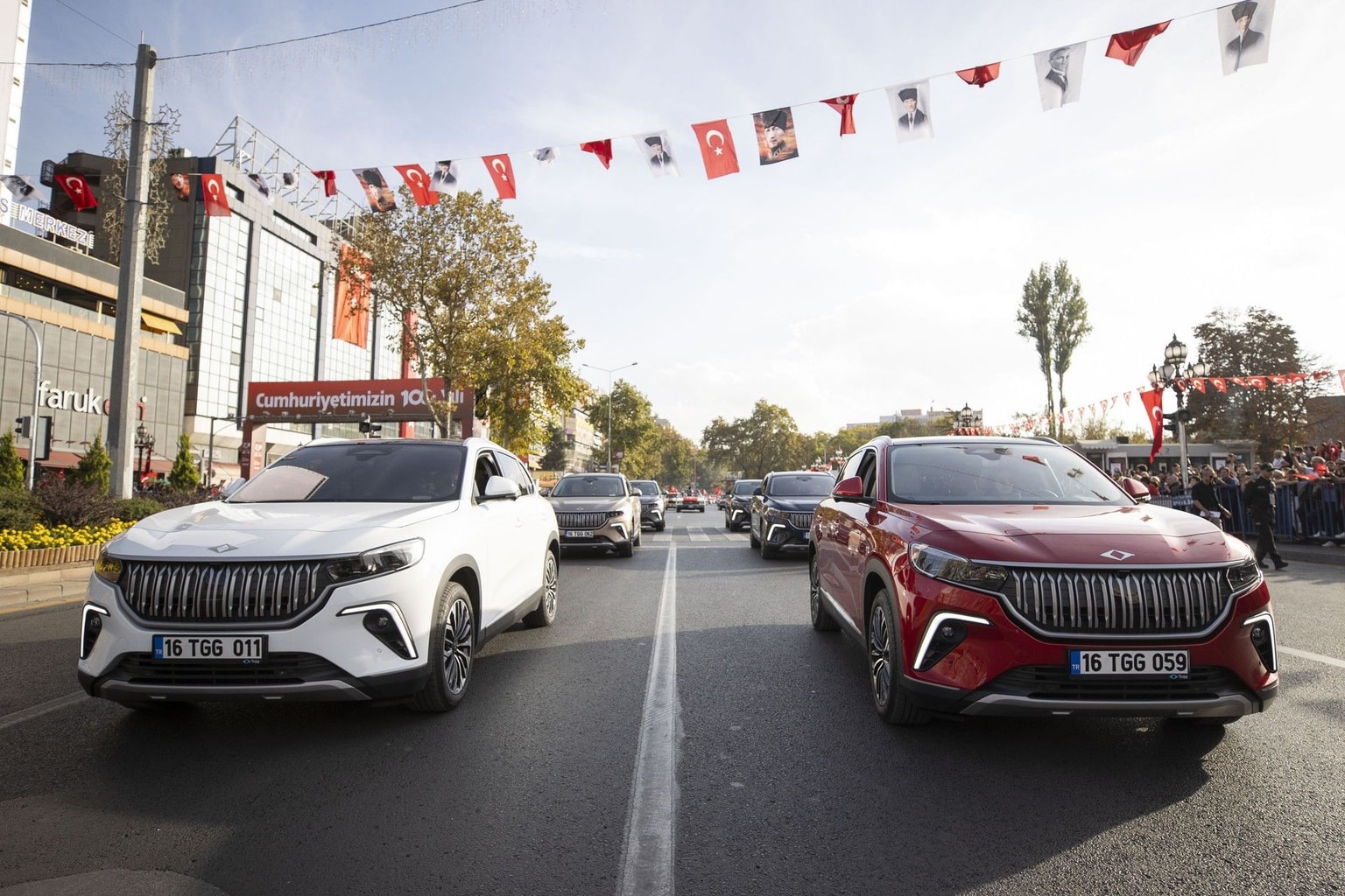 Due auto eleganti, una bianca e una rossa, vanno lungo una strada piena di bandiere turche e gente che guarda, sotto un cielo limpido.