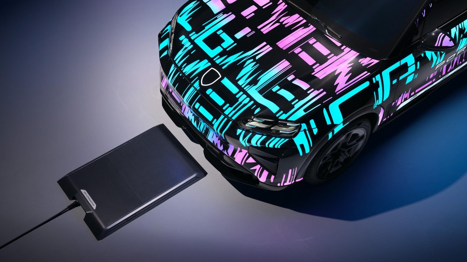A car with a vibrant geometric wrap in blue and pink is parked near a wireless charging pad, viewed from above.