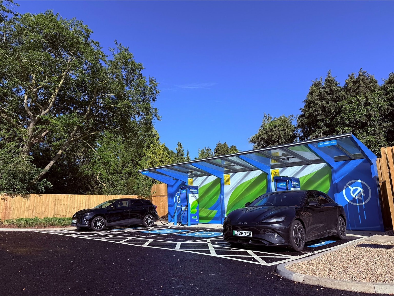Two black cars parked at a vibrant EV Power MFG electric vehicle charging station, surrounded by trees and clear blue skies.