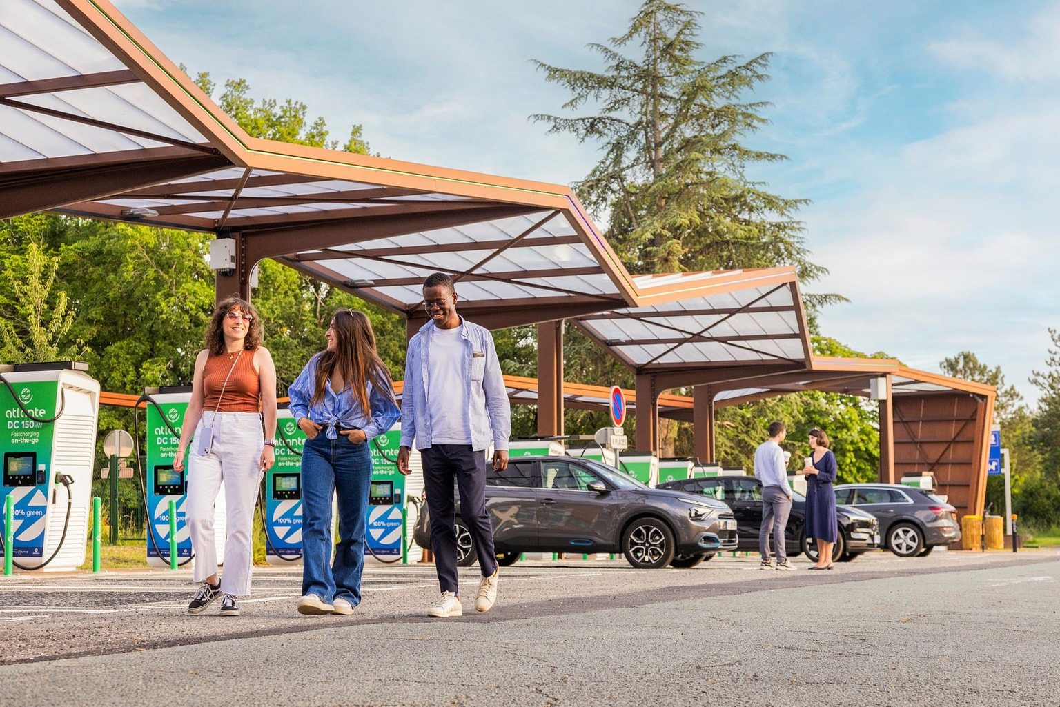 In una giornata di sole, alcune persone passeggiano accanto a una fila di stazioni di ricarica per veicoli elettrici Atlante, con auto parcheggiate sotto tettoie angolari dotate di pannelli solari.