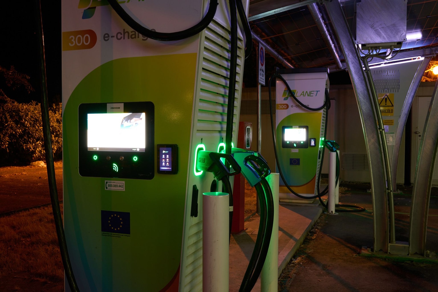 Stazione di ricarica per veicoli elettrici iPlanet di notte con schermi illuminati e indicatori di ricarica verdi sotto una tettoia.