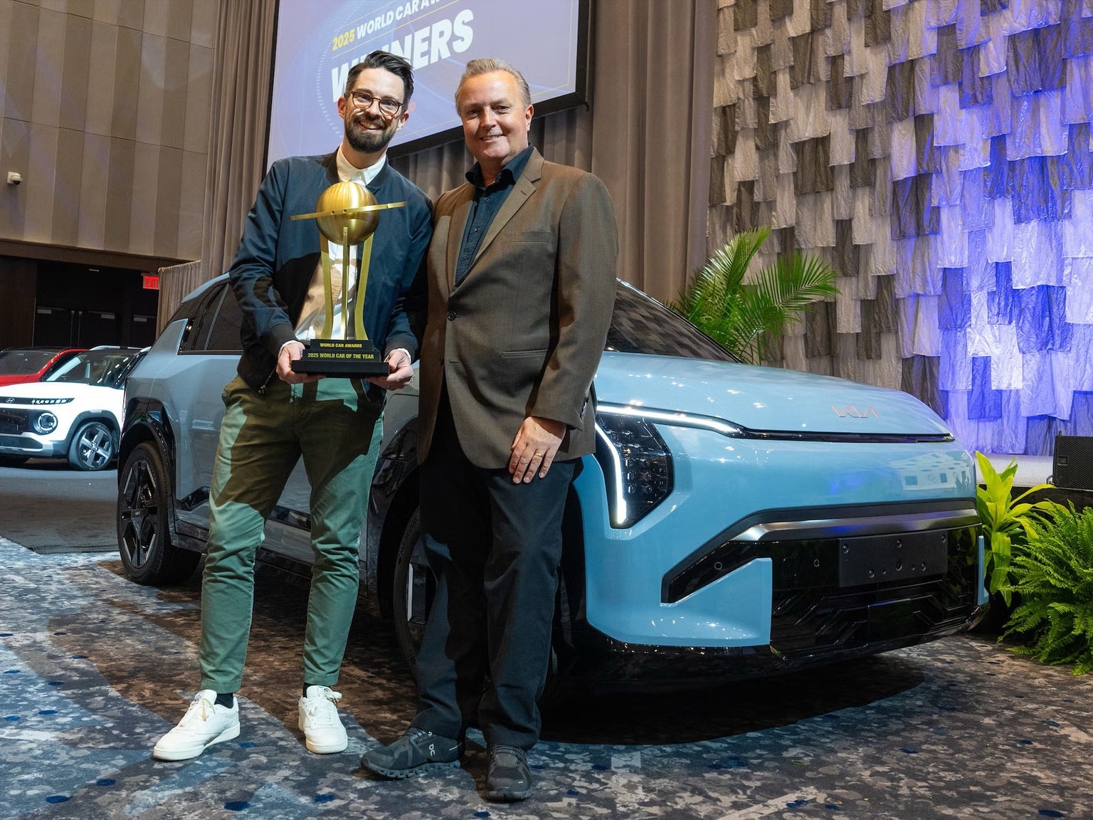 Two men stand beside a light blue KIA, holding a trophy. A backdrop reads "World Car Awards Winners." Due uomini sono in piedi accanto a una KIA azzurra, con in mano un trofeo. Sullo sfondo è scritto "Vincitori dei World Car Awards".