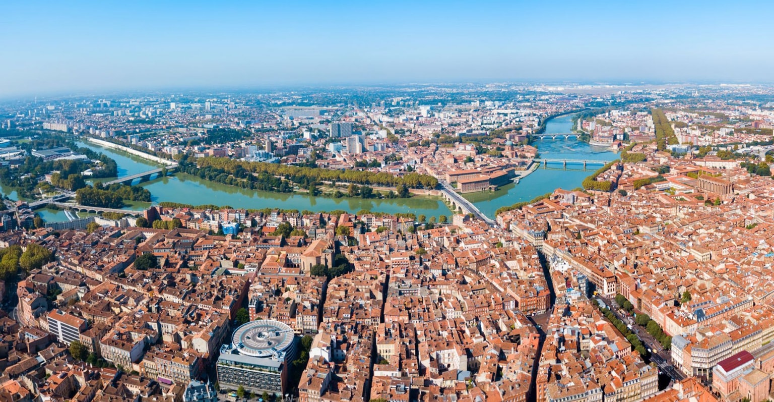 Vue aérienne de Toulouse avec ses toits de tuiles rouges, sa rivière sinueuse, ses multiples ponts et ses espaces verts sous un ciel bleu clair.