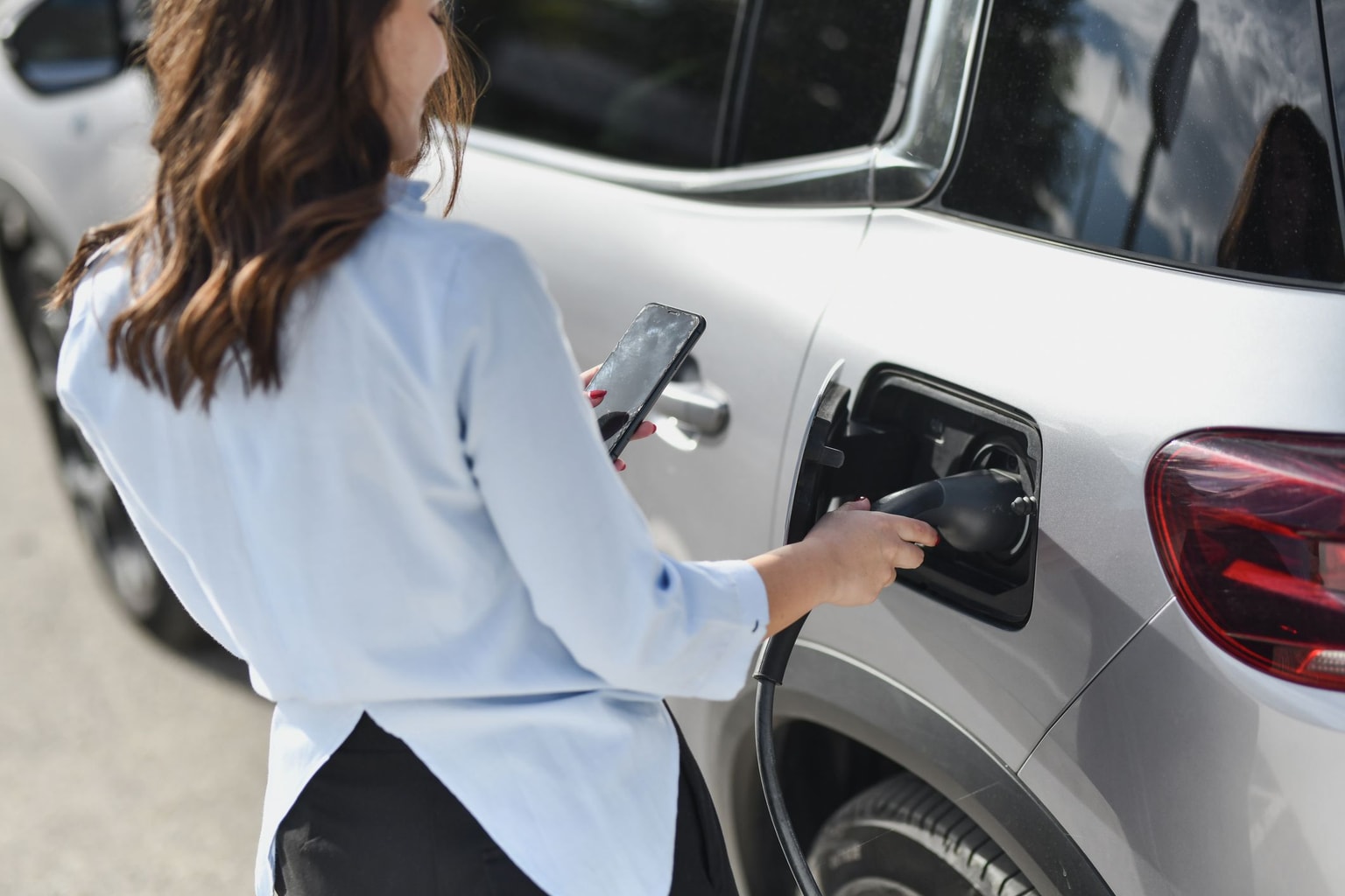 Eine Person in einem hellblauen Hemd schließt ein Ladegerät für Elektrofahrzeuge an einen silbernen SUV an, während sie ein Smartphone in der Hand hält.