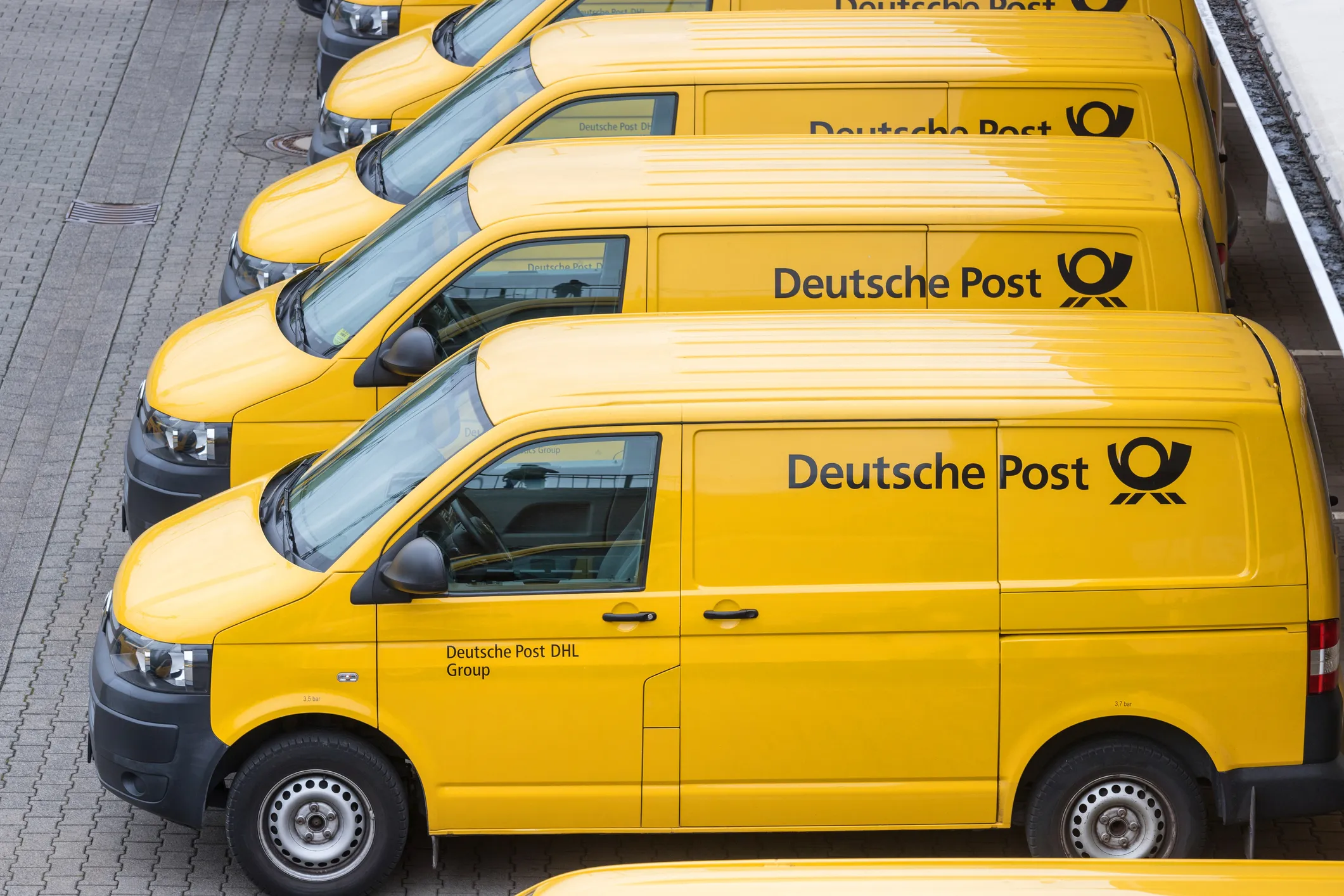 Eine Reihe gelber Zustellfahrzeuge der Deutschen Post, die auf einer gepflasterten Fläche stehen und das Firmenlogo sowie das Branding des Unternehmens tragen.