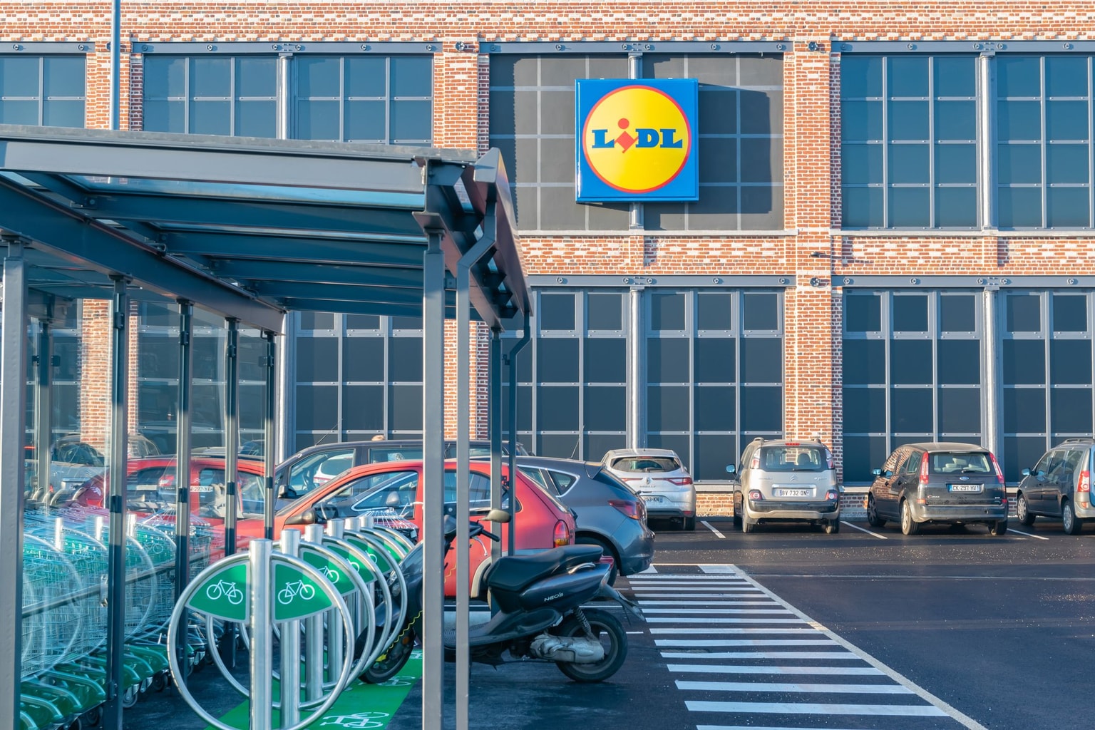 Parking avec voitures et vélos à l'extérieur d'un magasin Lidl, avec des murs en briques rouges et de grandes fenêtres sous un ciel bleu clair.