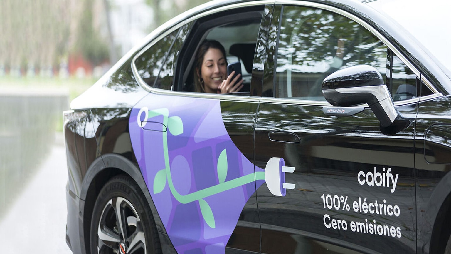 Una mujer sonriente utilizando su teléfono móvil dentro de un coche eléctrico negro con un diseño de enchufe verde. El coche muestra el mensaje «cabify 100 % eléctrico Cero emisiones».
