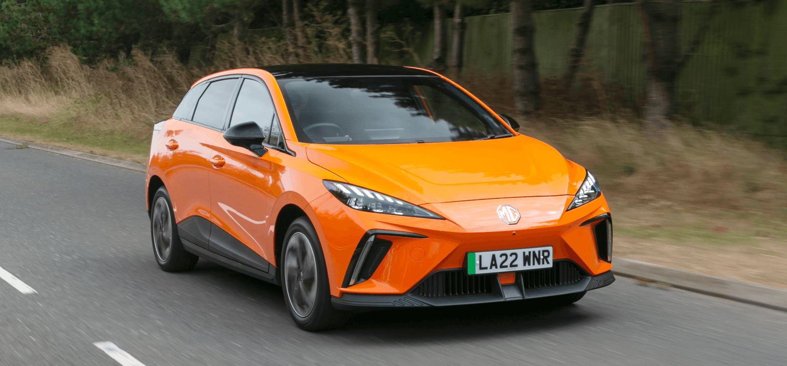 Un MG4 de color naranja brillante circulando por una carretera, rodeado de vegetación, con una matrícula visible "LA22 MNR".