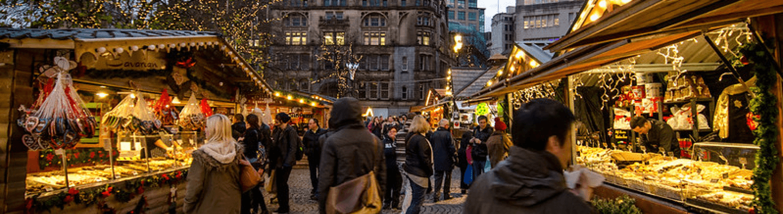 Persone che curiosano tra le bancarelle festive del mercatino di Natale di Manchester, illuminate da lucine, con cibo e decorazioni in mostra