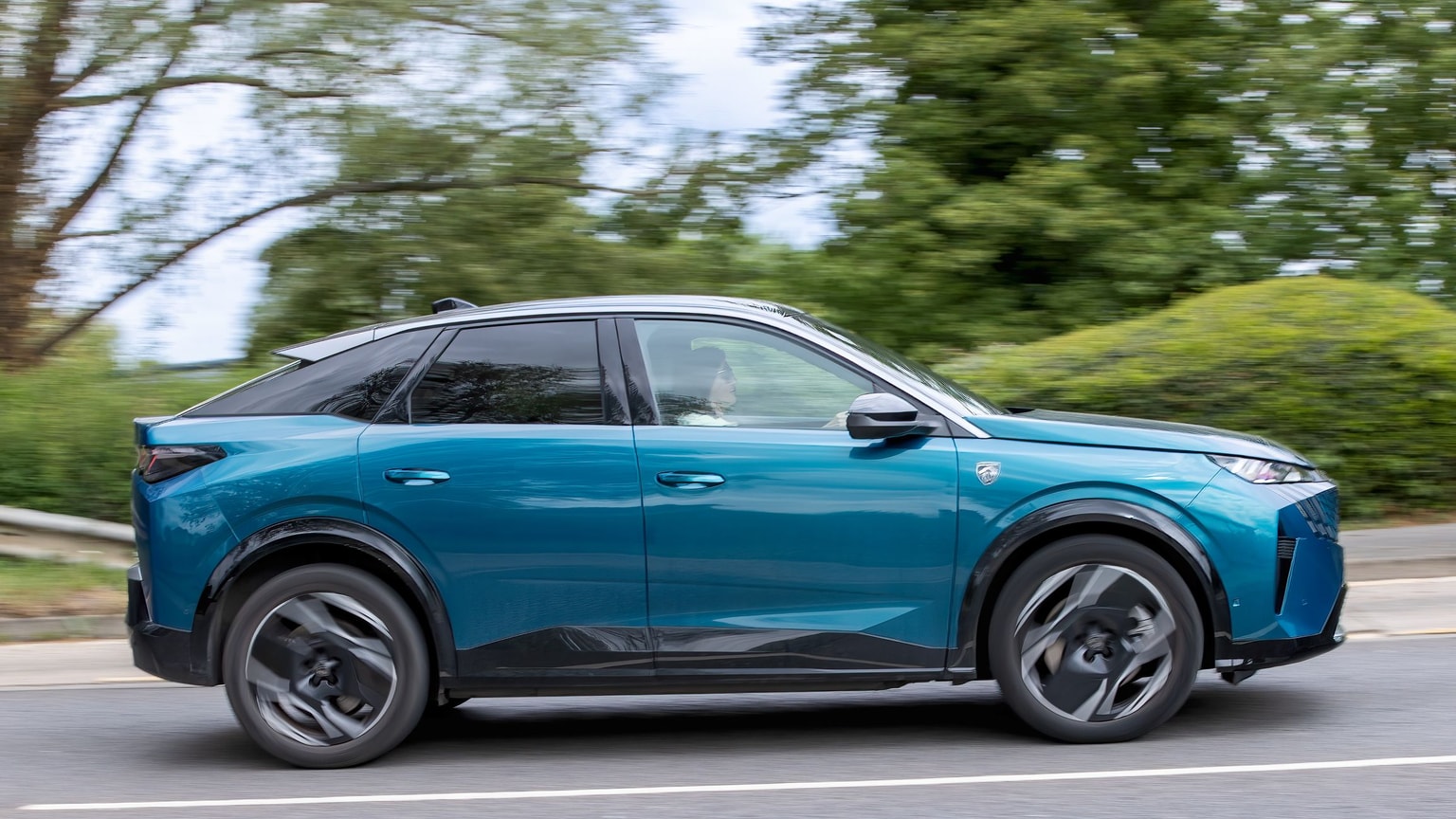 A blue Peugeot E-3008 SUV driving on a road with blurred green trees in the background, emphasizing motion.