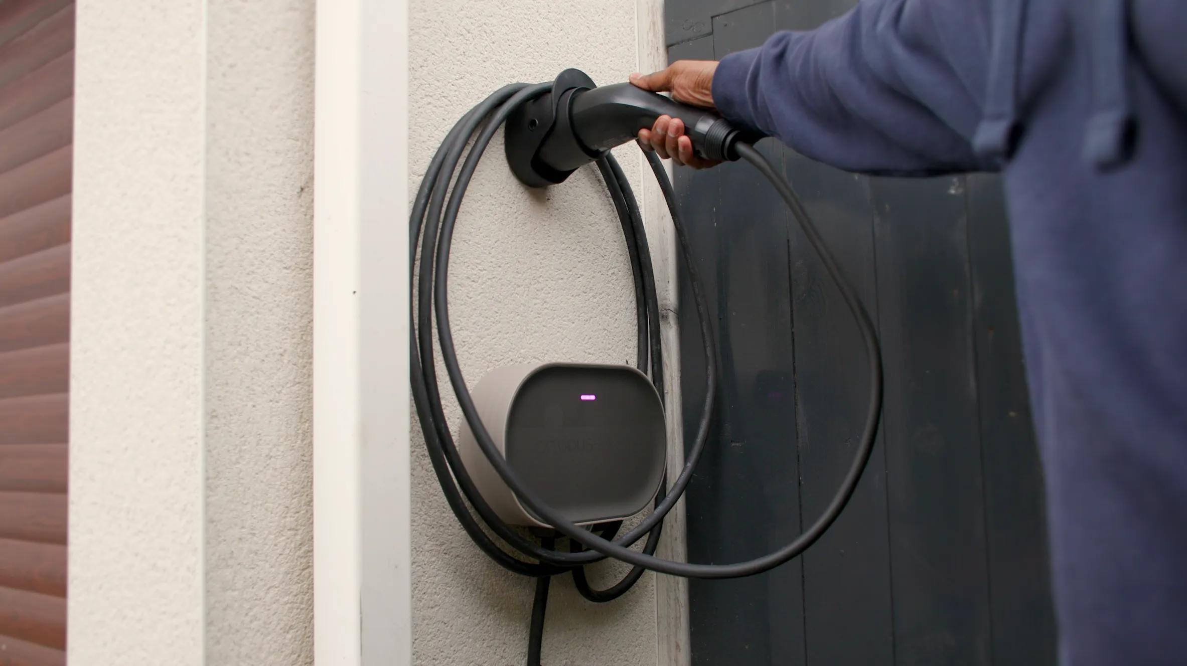 Une personne portant une veste bleue accroche un câble de recharge enroulé à une borne de recharge pour véhicules électriques fixée au mur.