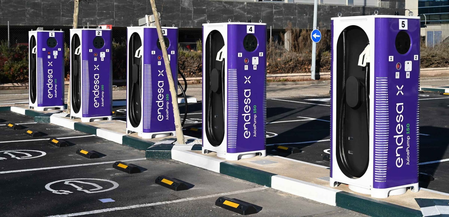Fila de estaciones de carga para vehículos eléctricos Endesa X de color morado en una zona de aparcamiento, cada una marcada con un número y símbolos EV en el suelo.