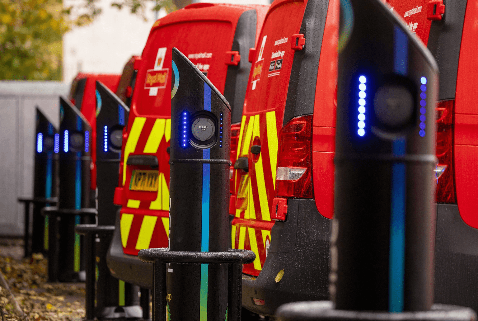Furgoni postali rossi in ricarica presso stazioni di ricarica elettrica, con luci LED blu sulle stazioni, in un ambiente esterno.