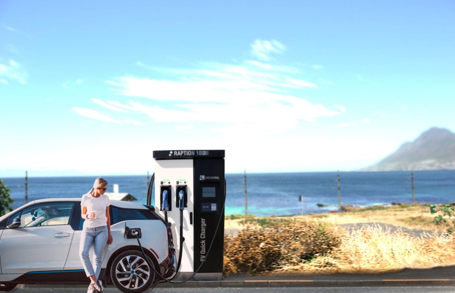 Una persona se apoya en un coche eléctrico que se carga en una estación junto al océano, con montañas y un cielo azul claro de fondo en España.