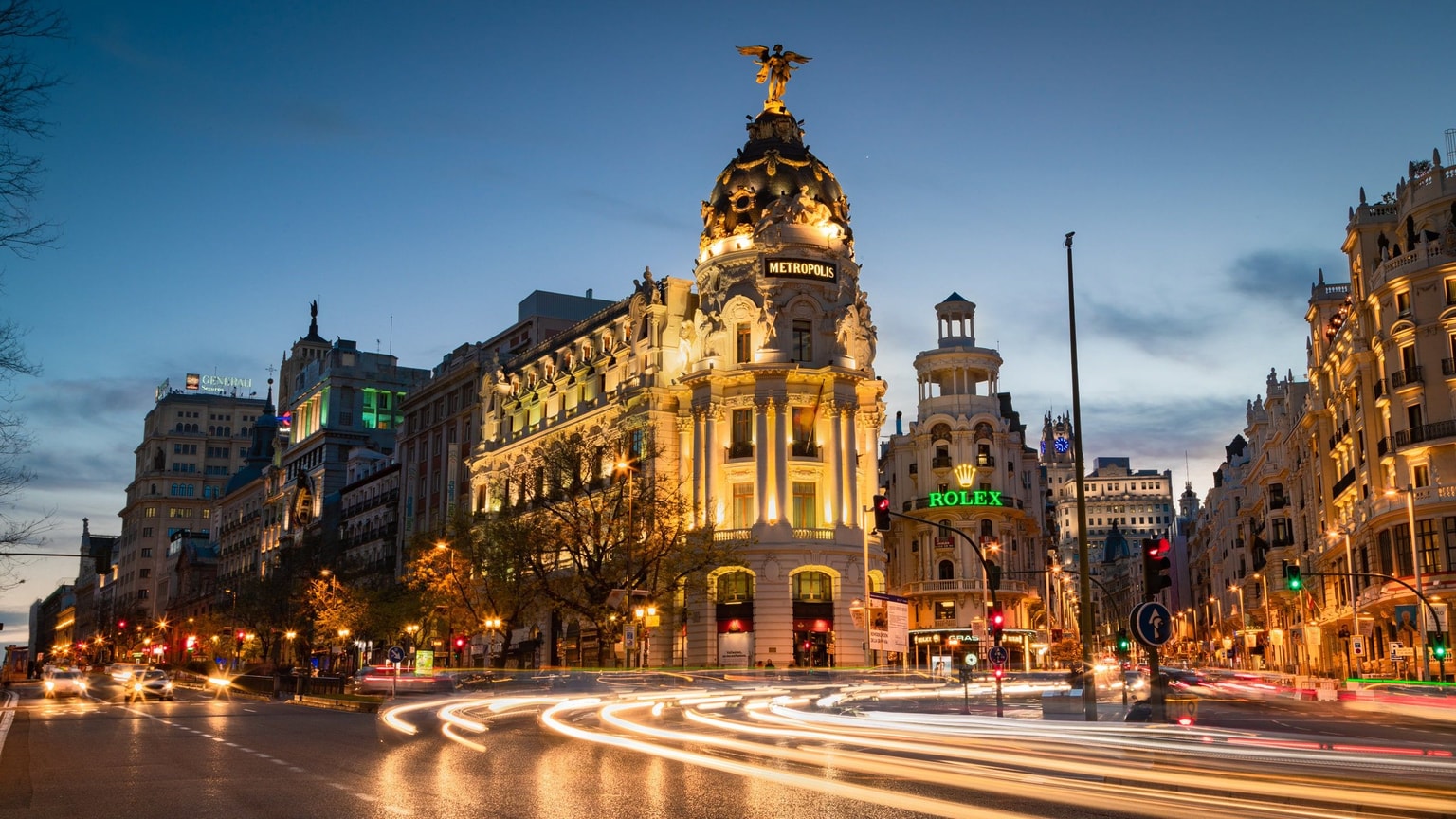 Vista nocturna del Edificio Metrópolis de Madrid, con estelas de luz del tráfico en la Gran Vía y arquitectura circundante iluminada.
