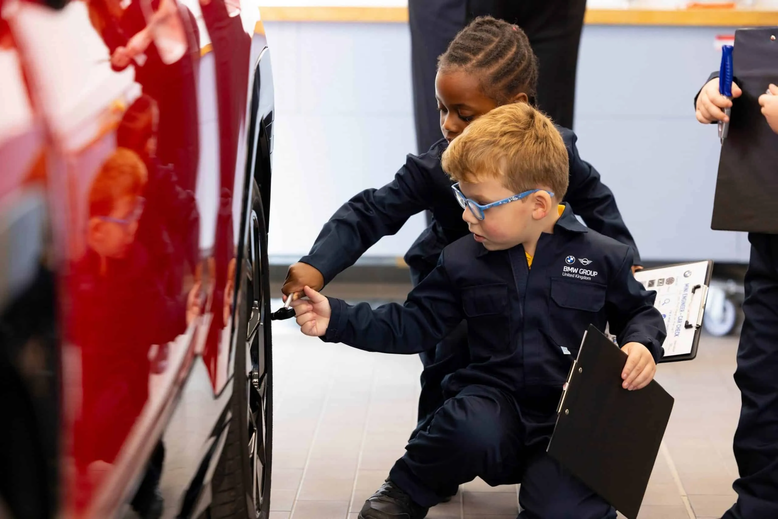 Zwei Kinder in dunkelblauen Uniformen untersuchen in einer Werkstatt einen Autoreifen und halten Klemmbretter in der Hand.