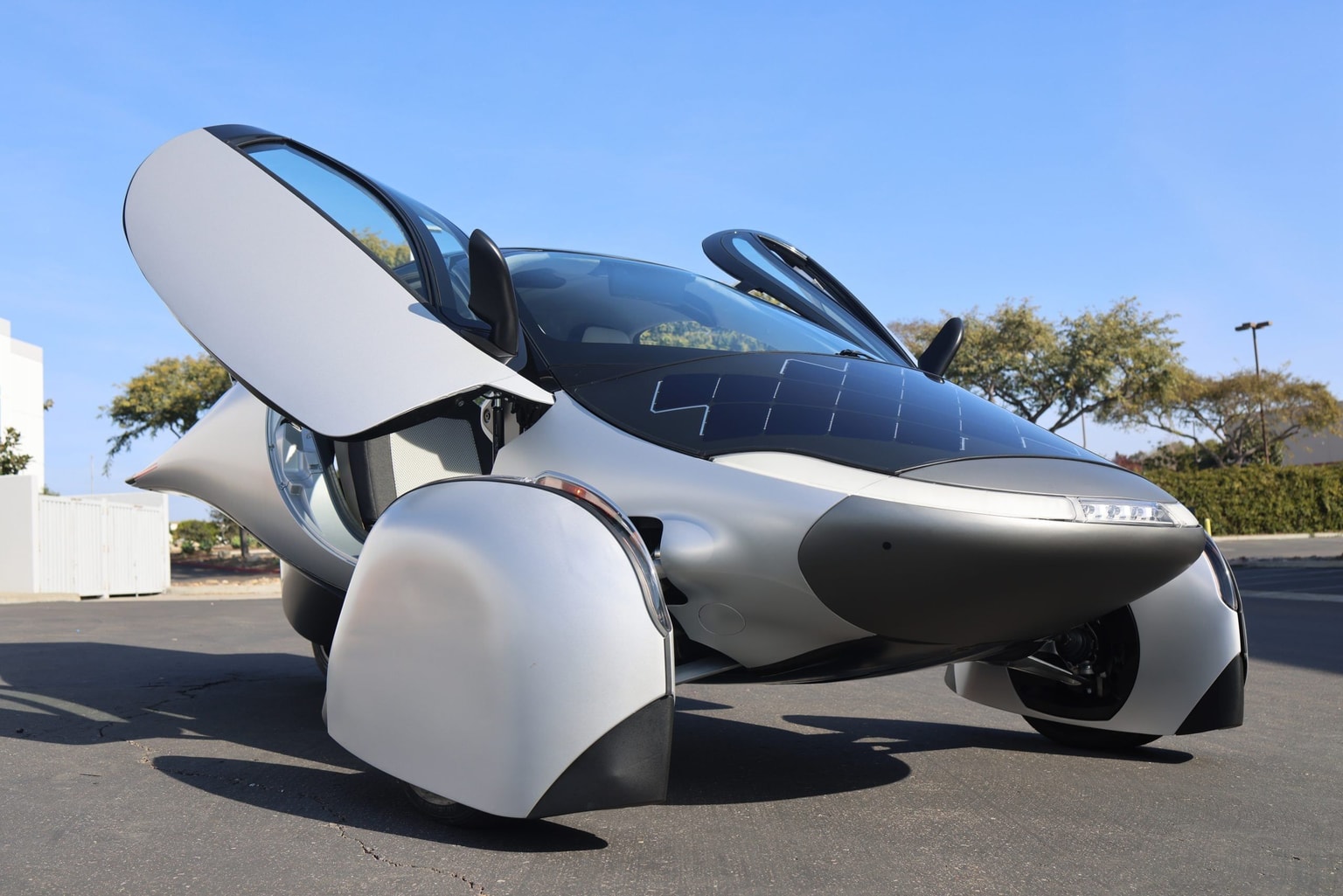 Vehículo eléctrico futurista plateado con puertas abiertas tipo mariposa, paneles solares en el capó y un elegante diseño aerodinámico, estacionado al aire libre.