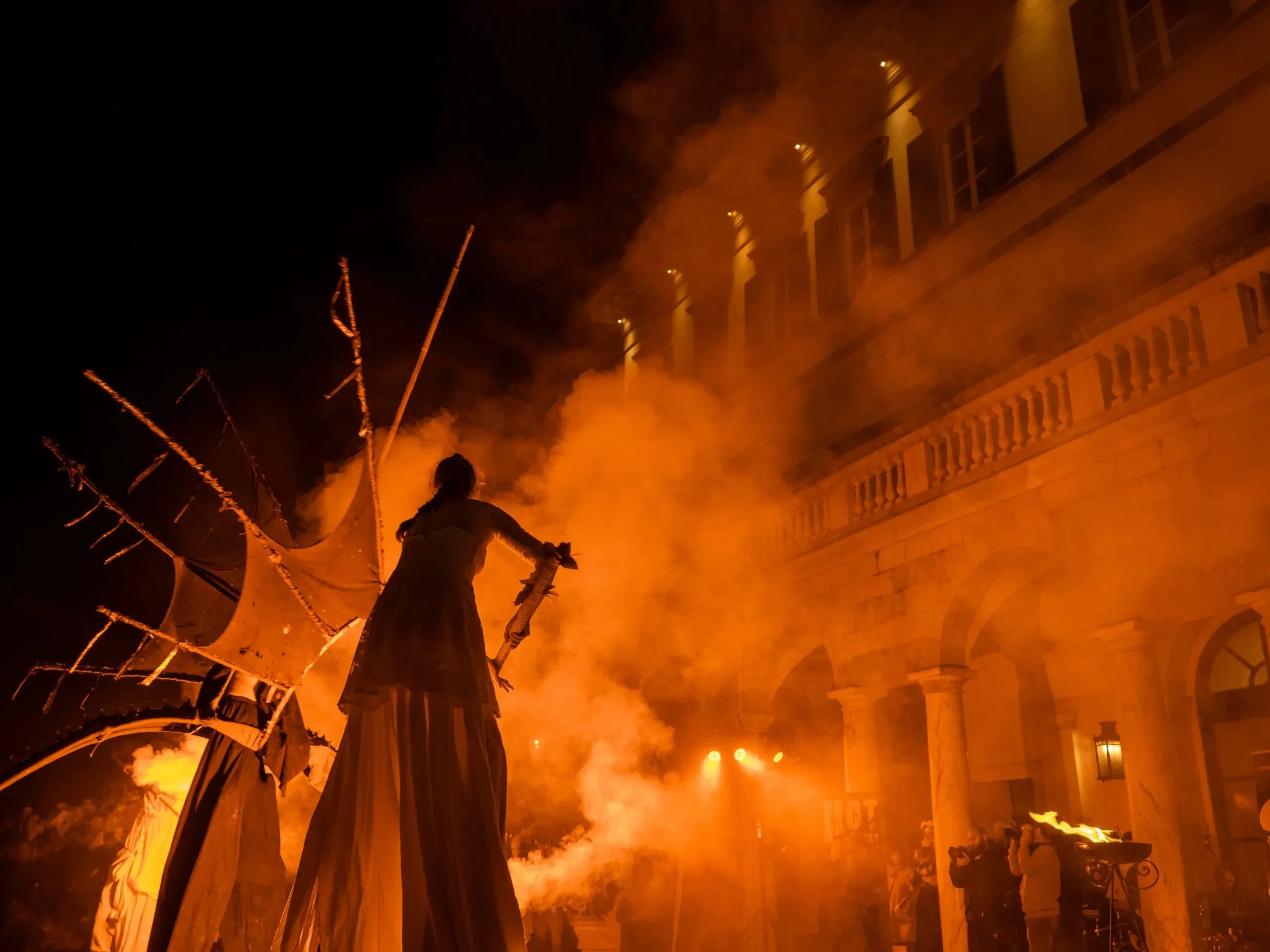 Una persona sui trampoli, vestita con una lunga tunica, sta in piedi tra fumi arancioni, illuminata dalle luci di scena, di fronte a un edificio storico durante la notte.