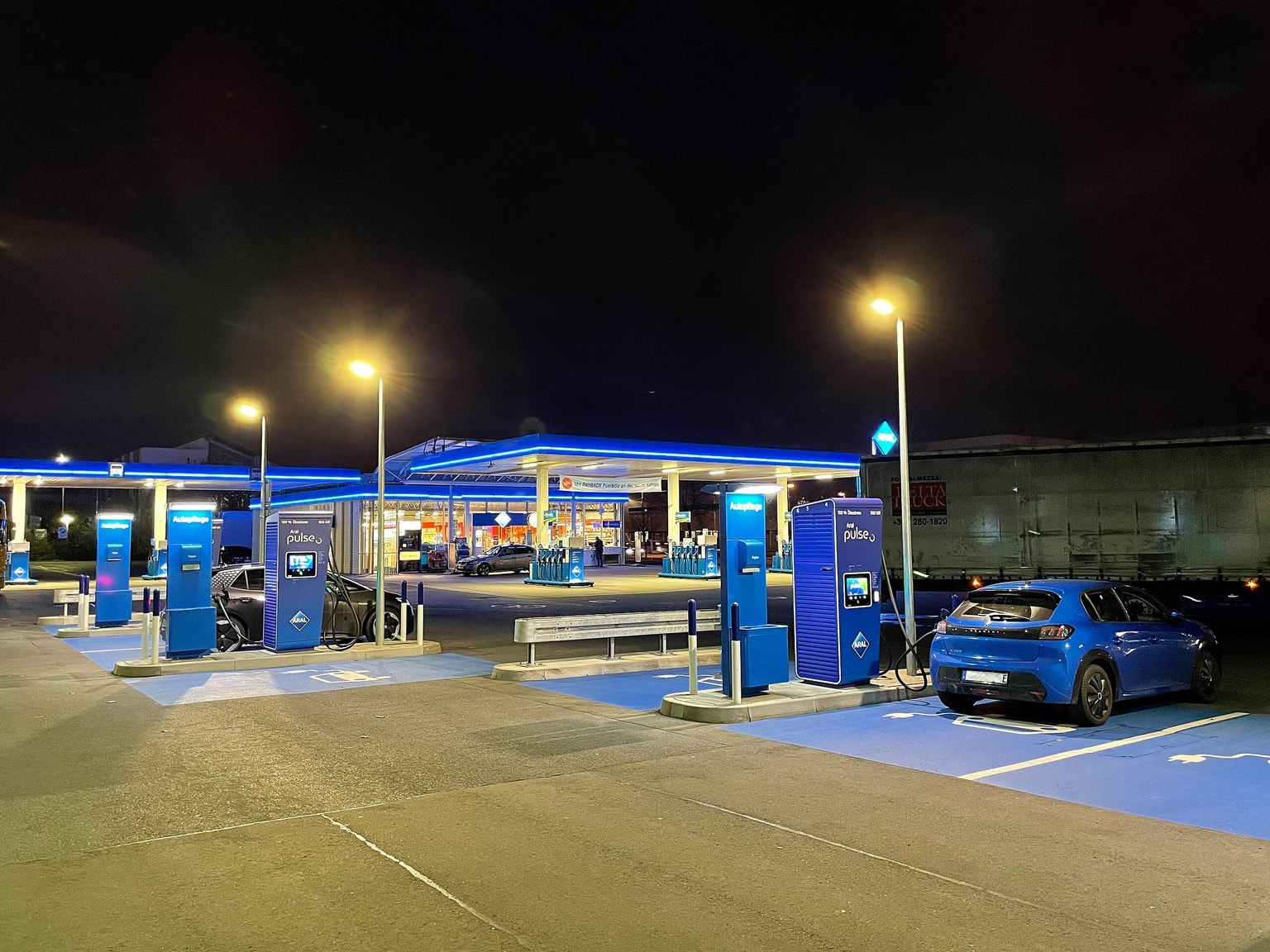 Elektroauto-Ladestation bei Nacht mit einem geparkten blauen Auto, beleuchtet von Straßenlaternen, und einem Supermarkt im Hintergrund.