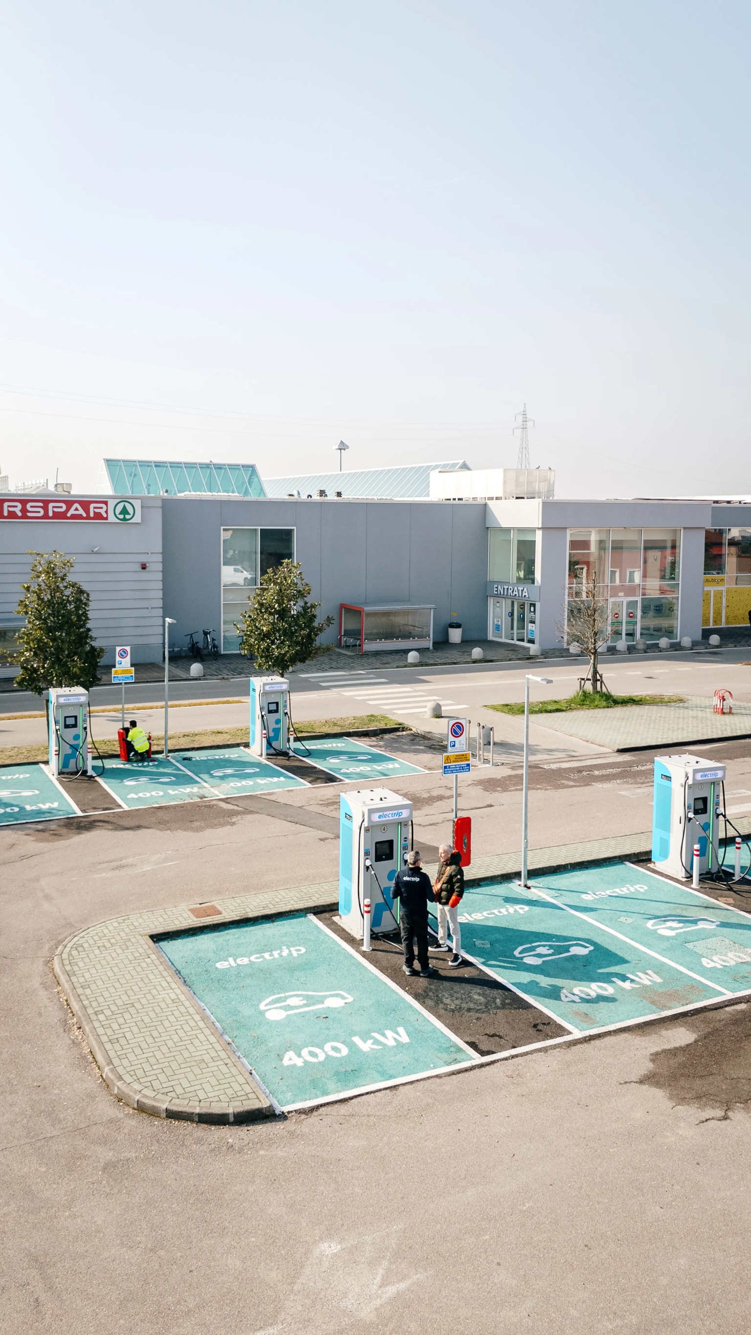 Stazione di ricarica per auto elettriche Electrip con più punti di ricarica all'esterno di un negozio, con persone che interagiscono vicino a un caricabatterie.