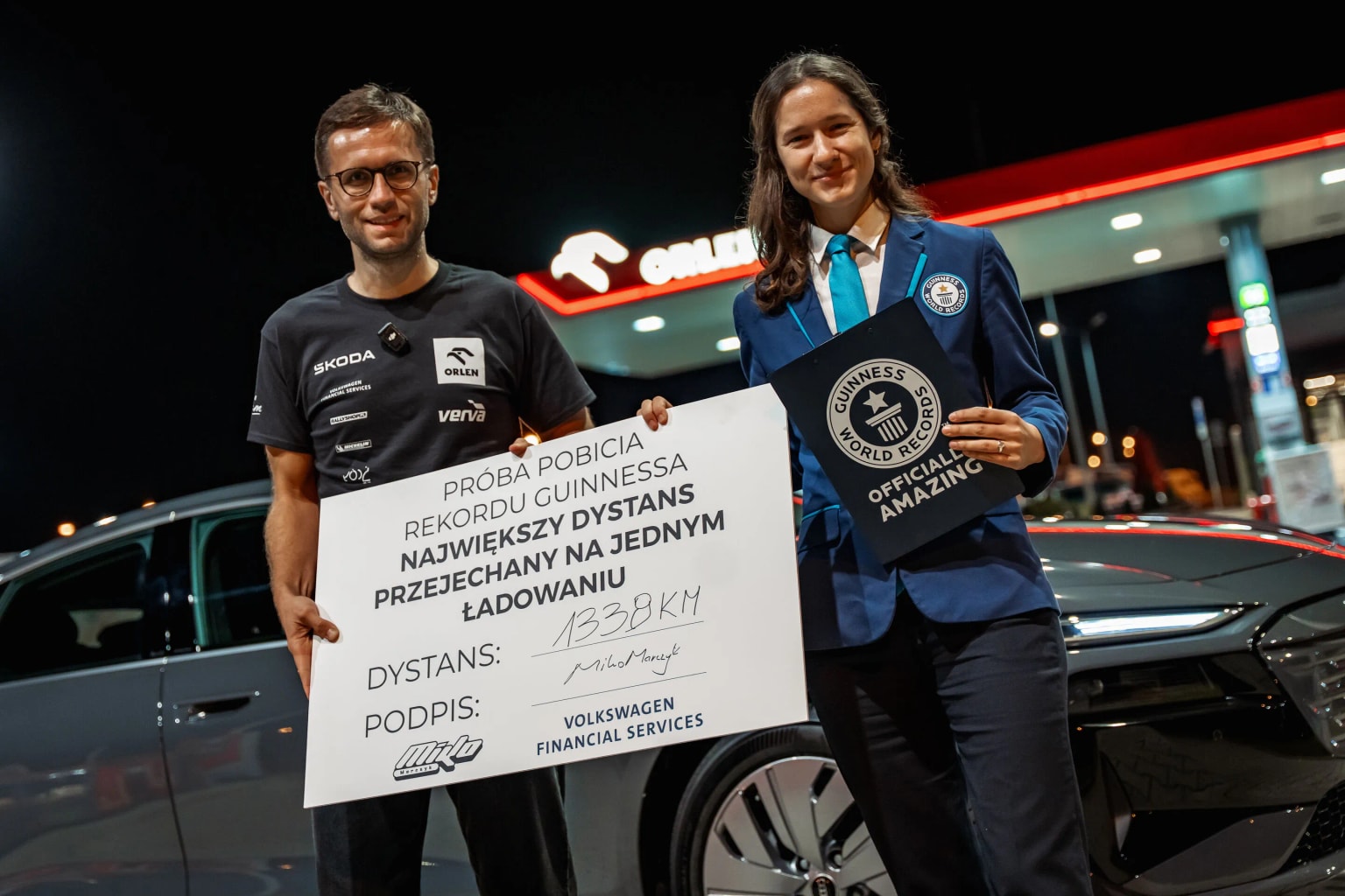 Ein Mann und eine Frau stehen an einer Tankstelle. Der Mann hält ein Schild mit Einzelheiten zum Rekord. Die Frau trägt einen blauen Anzug und hält eine Guinness-World-Records-Plakette.
