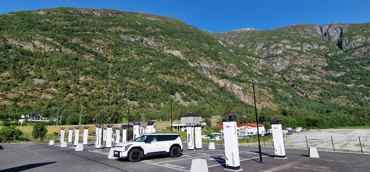 A white car parked at a IONITY charging station with multiple charging units, set against a backdrop of lush green mountains.