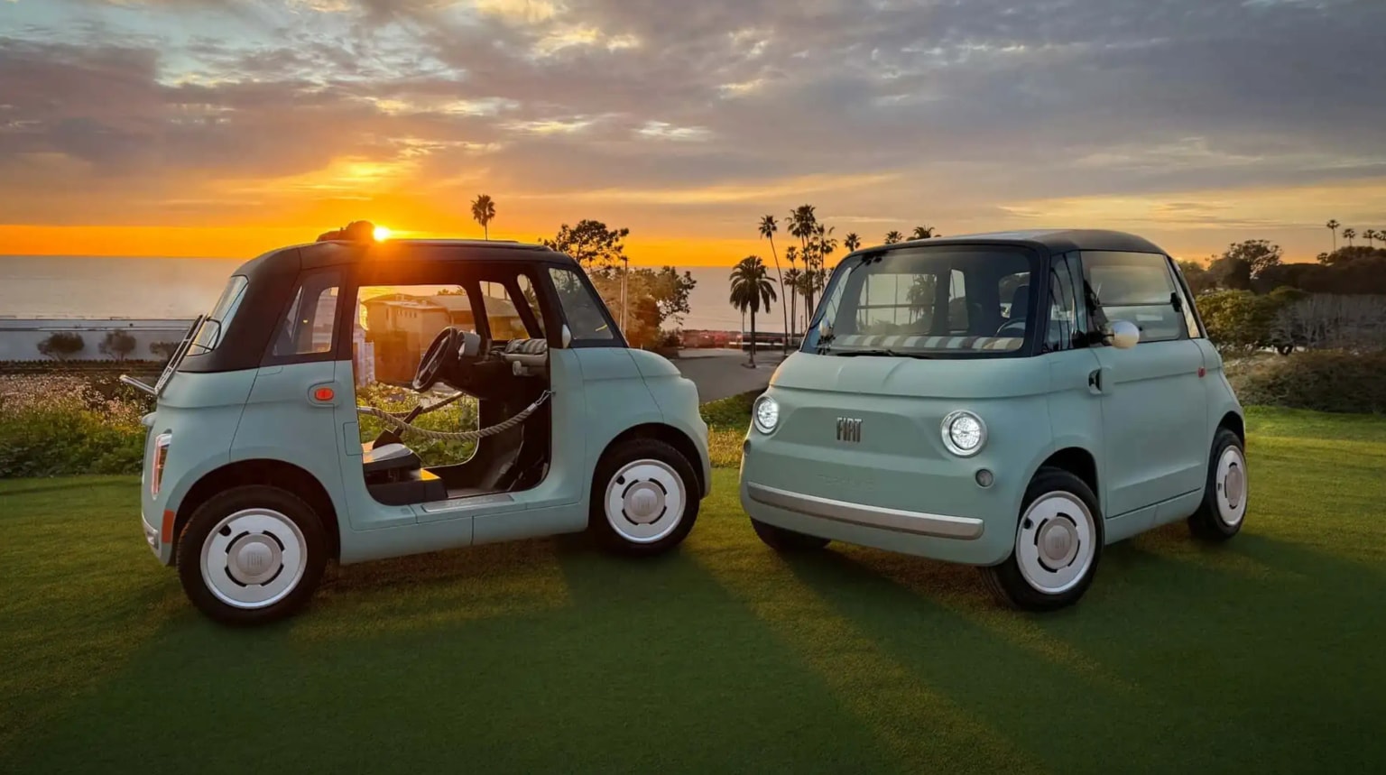 Deux petites voitures Fiat aux couleurs pastel garées sur l'herbe, avec un coucher de soleil pittoresque et des palmiers en arrière-plan.