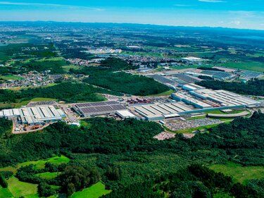 Vue aérienne d'un vaste complexe industriel avec des bâtiments d'usine au toit blanc au milieu de forêts verdoyantes et de terres agricoles.