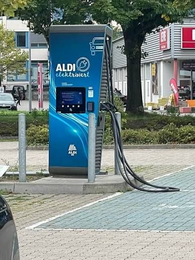 A blue Aldi electric vehicle charging station is positioned in a parking lot, surrounded by a few bollards, with buildings in the background.