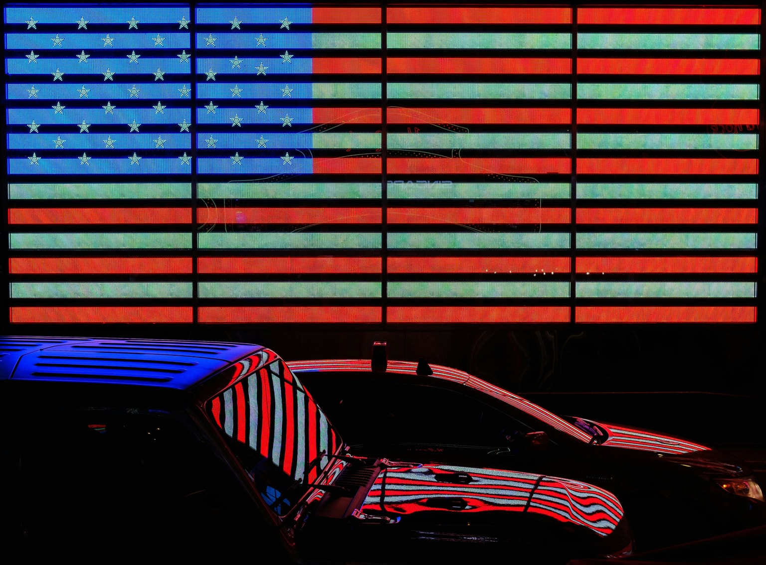 Two cars reflect a brightly lit digital American flag display at night.