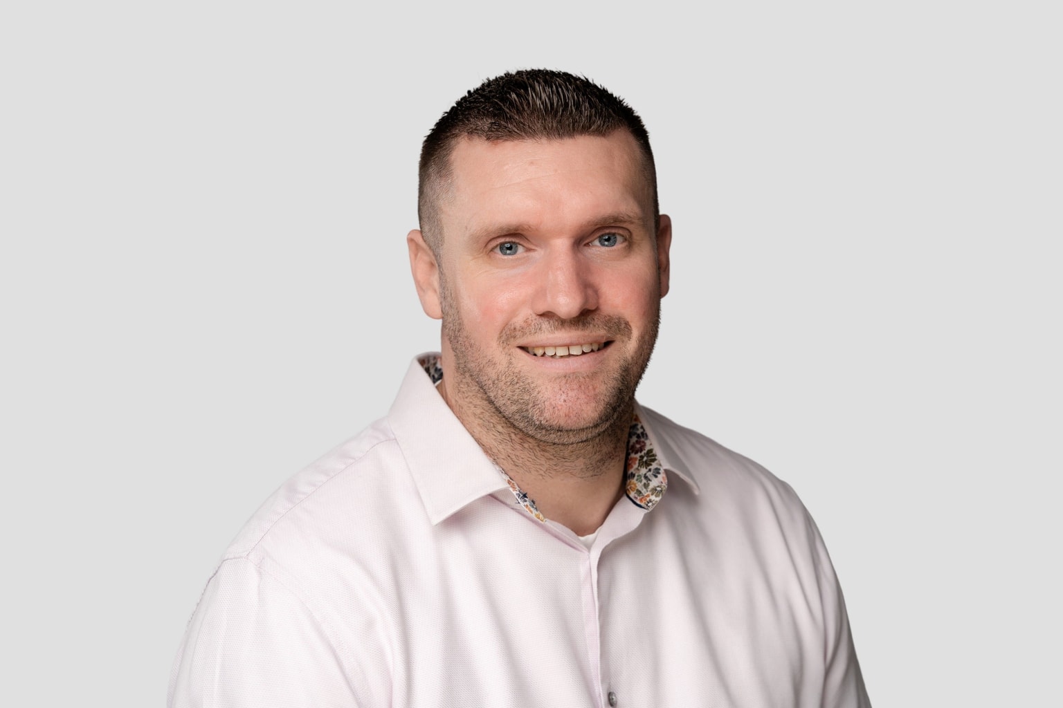 Smiling man in a light pink shirt with a floral collar detail, set against a plain gray background.